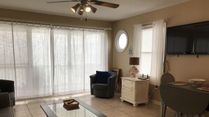 A living room with a glass-top coffee table, a sofa, a chair, a small white cabinet, a dining table with two chairs, a ceiling fan, and a TV mounted on the wall.