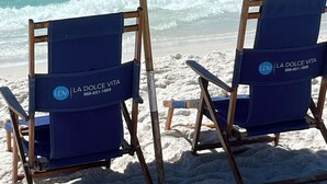 Two beach chairs with 'La Dolce Vita' branding and a large blue umbrella on a sandy beach with waves in the background.