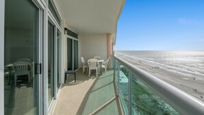 A balcony with a glass door, a table, and chairs, overlooking a beach. - Lovely Condo, Direct Oceanfront Views, Pools | Crescent Keyes - 1209 (North Myrtle Beach)