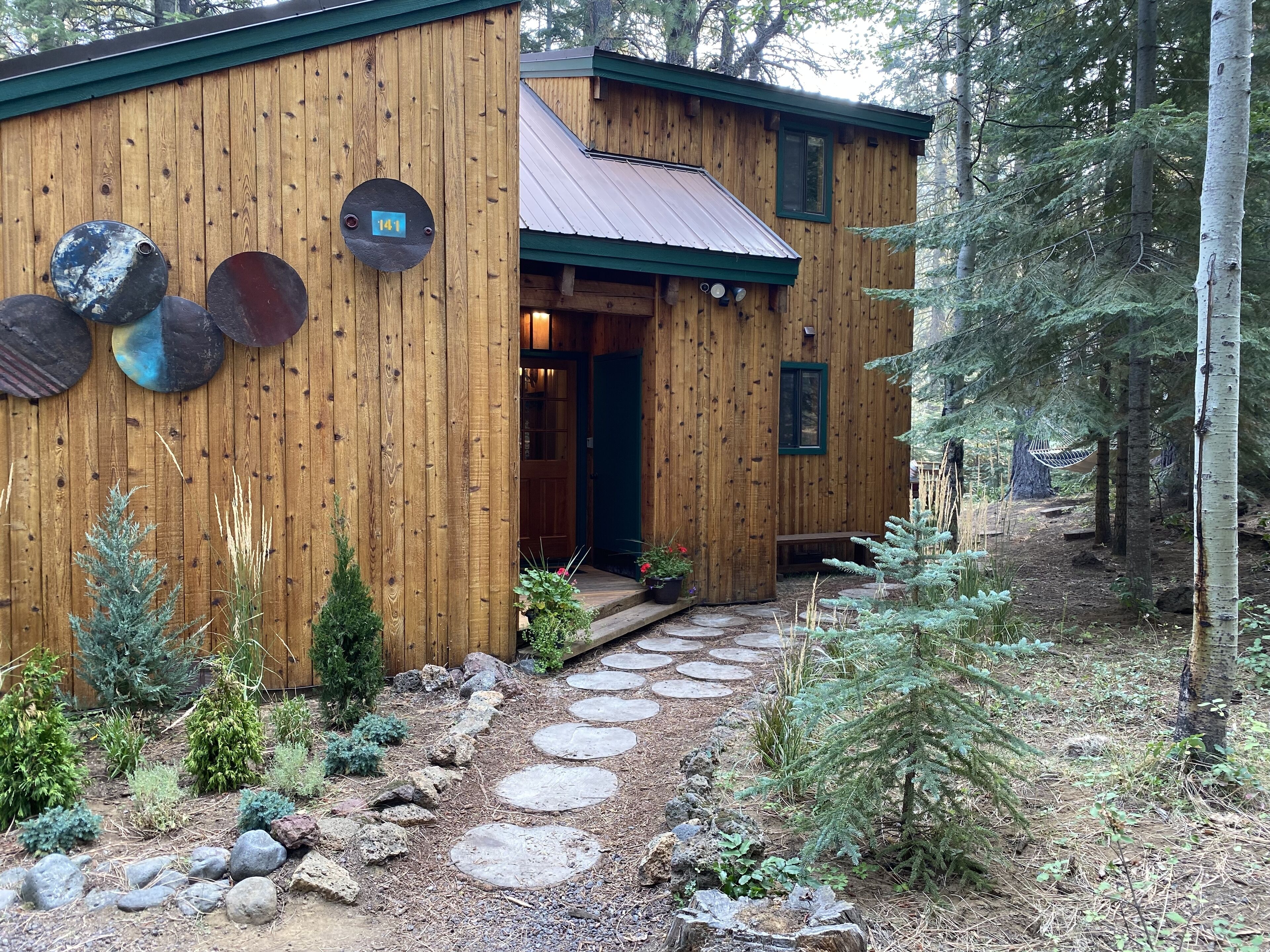 A wooden cabin with a stone pathway leading to the entrance, surrounded by a forest.