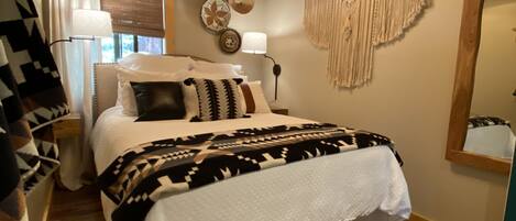 A cozy bedroom with a wooden ceiling, a bed with patterned bedding, a hanging macrame wall decoration, and a large mirror.