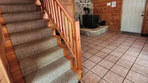 A wooden staircase with carpeted steps and a wooden handrail, leading to a room with a stone wall, a black stove, and a white door. - 2400sf Secluded Family Mountain House 16 Acres- 2 min to Jiminy Peak- near Lenox (Hancock)