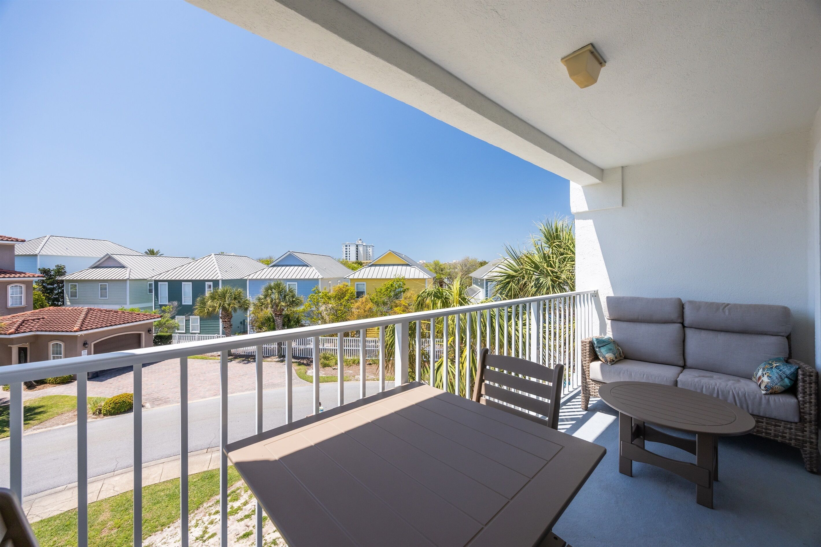 A balcony with a sofa and table, overlooking a residential area with houses and a clear sky.