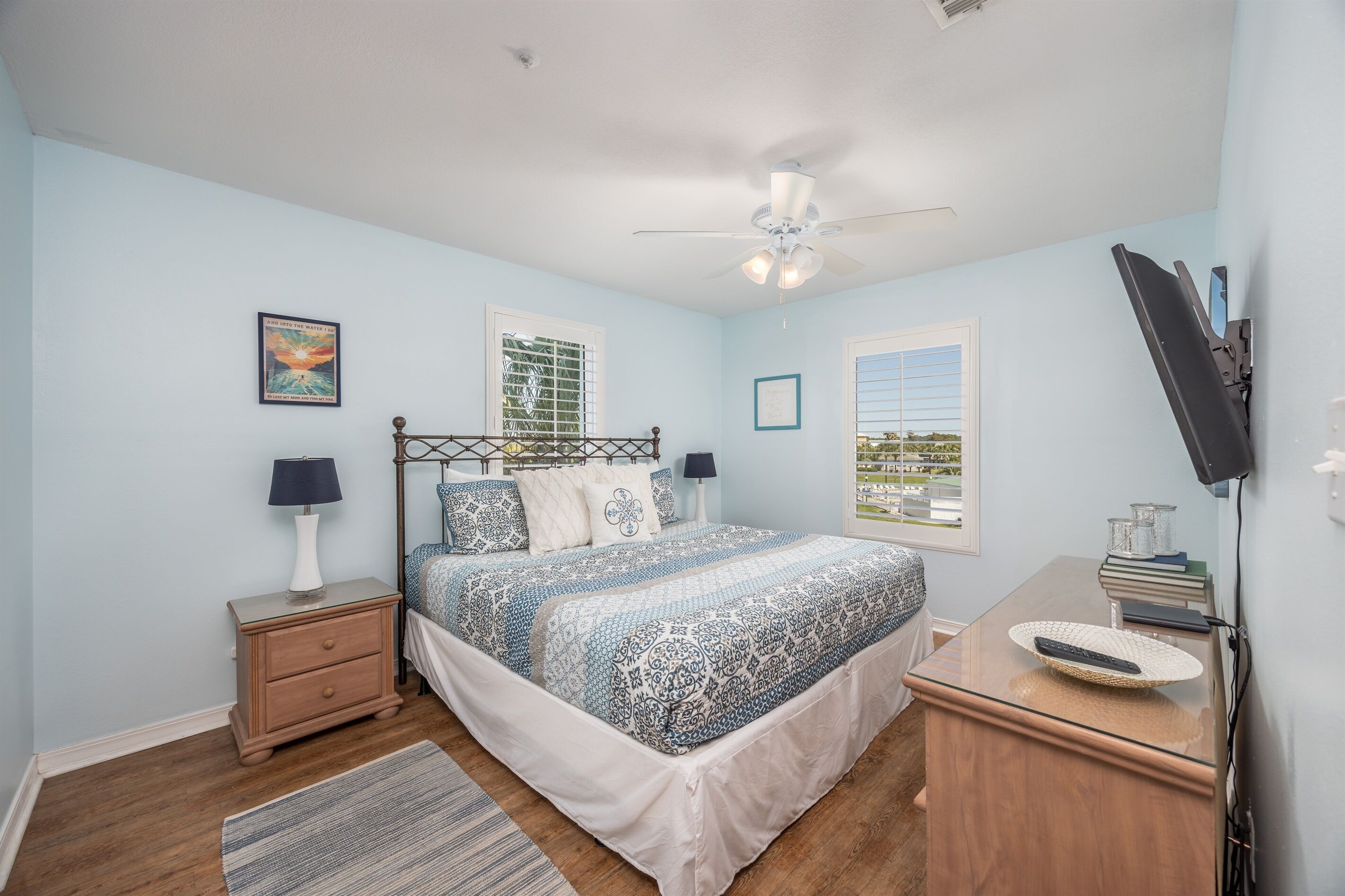 A bedroom with a bed, nightstands, a desk, a television, and windows.