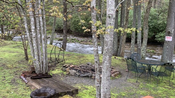 A rustic campsite with a picnic table, a bench, and a metal chair near a stream. - Cozy Little Cabin Located Along The Banks Of A Beautiful Trout Stream (Lansing)