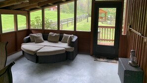 A cozy seating area with a cushioned bench and a small table, large windows offering a view of a grassy yard and a wooden deck. - Cozy Little Cabin Located Along The Banks Of A Beautiful Trout Stream (Lansing)