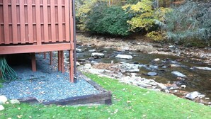 A wooden structure with a sloped roof, possibly a cabin or shed, adjacent to a rocky stream with a grassy foreground and trees in the background. - Cozy Little Cabin Located Along The Banks Of A Beautiful Trout Stream (Lansing)