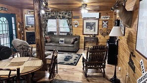 A cozy cabin interior with a wooden interior, a fireplace, a sofa, a dining table with chairs, and a mounted deer head. - Cozy Little Cabin Located Along The Banks Of A Beautiful Trout Stream (Lansing)