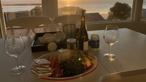 Mesa preparada para una comida con vista al atardecer, incluyendo una botella de vino, queso, galletas, uvas y una fuente de fruta.