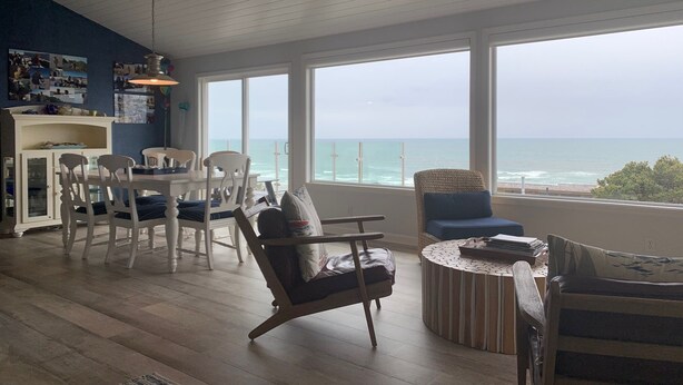 Un salón con vistas al océano, suelo de madera y una zona de comedor con sillas blancas y una mesa.