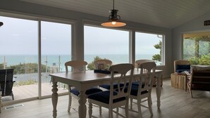 Un comedor con mesa y sillas, ventanas amplias con vistas al mar y una lámpara colgante.