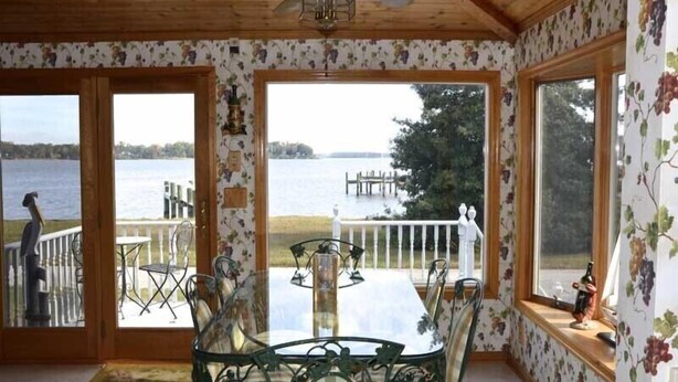 A room with a wooden ceiling, a ceiling fan, a table with chairs, and large windows offering a view of a lake and dock.