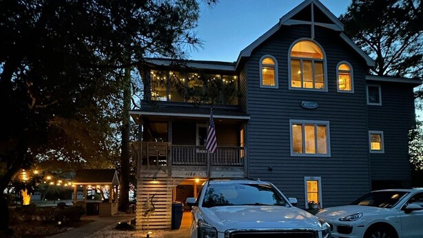 A two-story house with a balcony, an American flag, and a car parked in front. - BLUE HAVEN-COROLLA-OBX (Corolla)