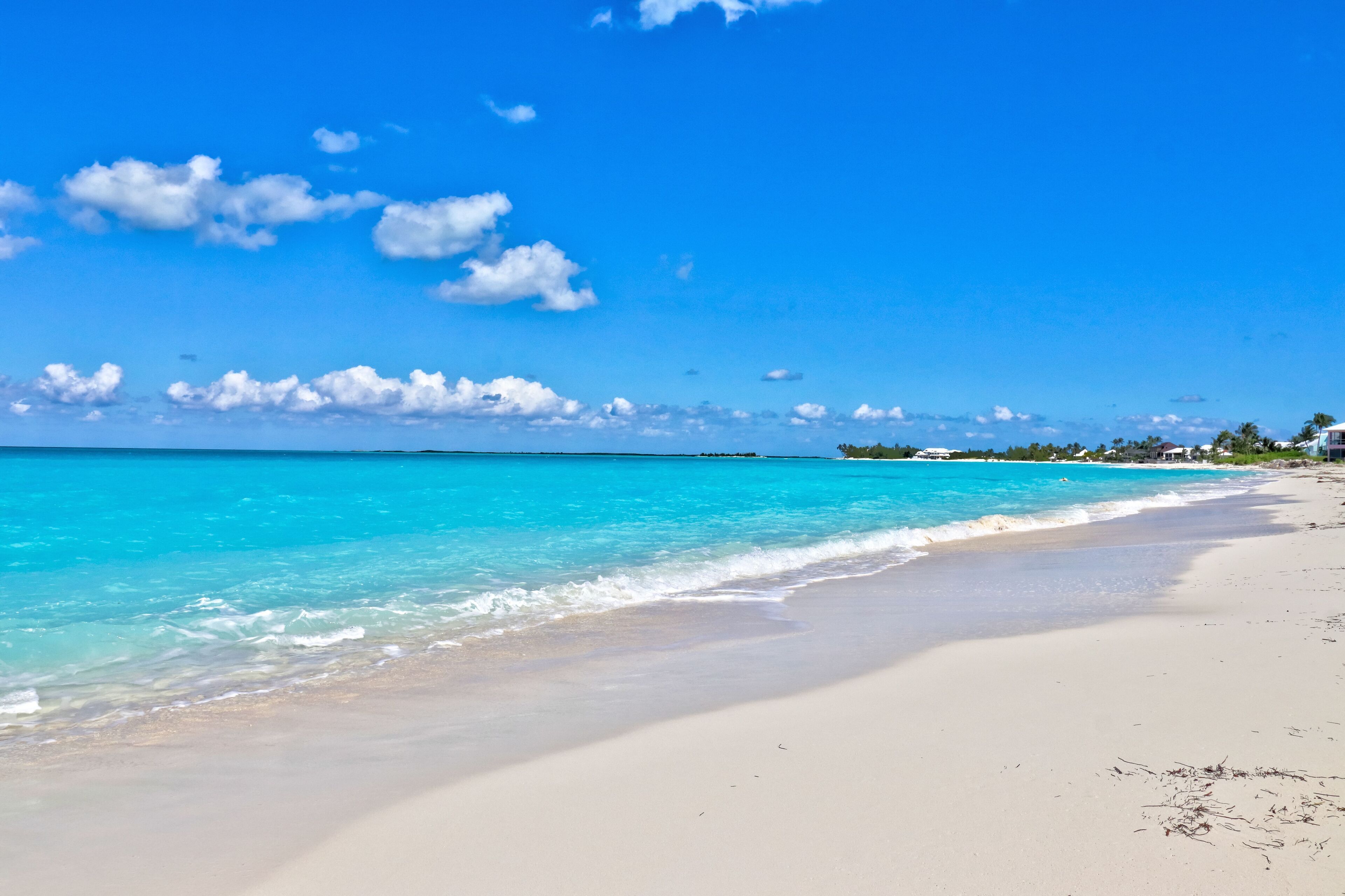 A beach with clear turquoise water, white sand, and a few buildings in the distance.