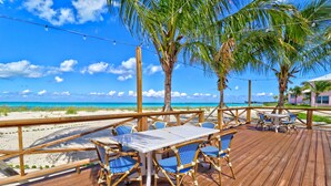 Terraza de madera con una mesa y sillas, con vista a una playa de aguas azules y palmeras.