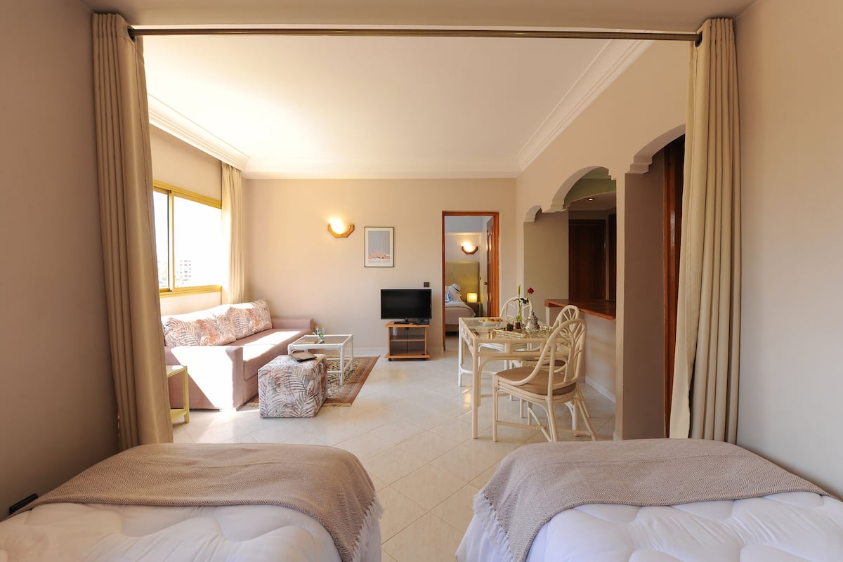 A hotel room with two beds, a sofa, a TV, a dining table, and a mirror.