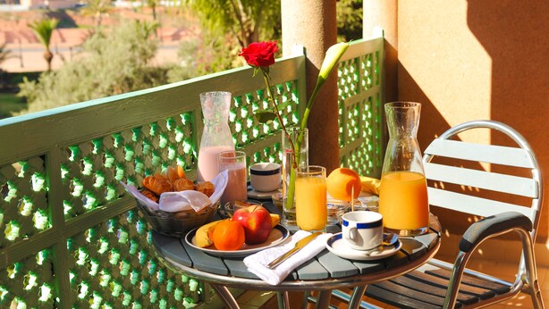 Una mesa dispuesta con alimentos para el desayuno, incluyendo frutas, una rosa y bebidas, en un balcón con vistas a edificios y árboles.
