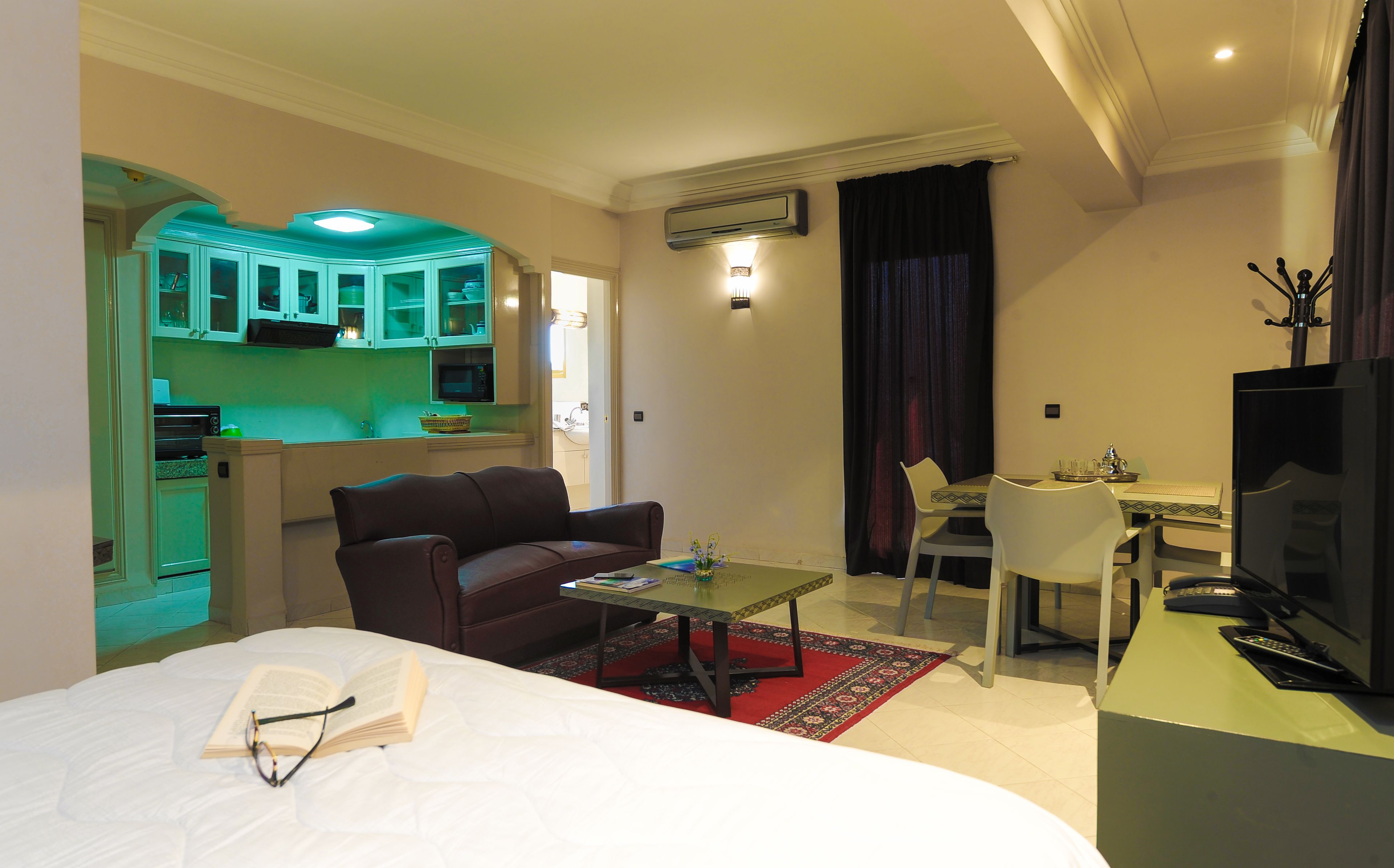Una habitación de hotel compacta con cama, un pequeño sofá, una mesa de comedor con sillas, un rincón de cocina y un televisor.
