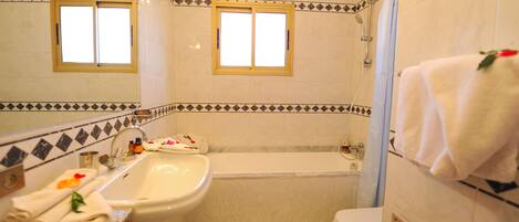 Ein Badezimmer mit Badewanne, Waschbecken und Toilette. Zwei Fenster und eine Duschvorhang sind vorhanden.