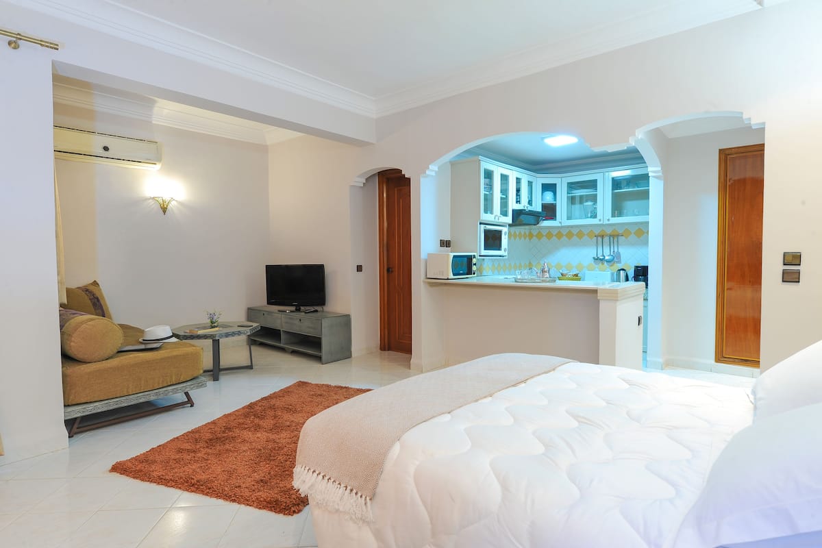 A bedroom with a bed, a sofa, a TV, a small table, and a kitchen area visible through an archway.