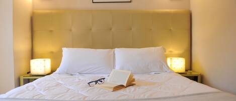 A neatly made bed with white linens, a book, and glasses on it. There are two bedside lamps and a framed picture on the wall above the bed.