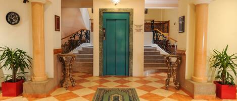 Un vestíbulo con suelo a cuadros, un ascensor verde y una escalera con barandillas decoradas.
