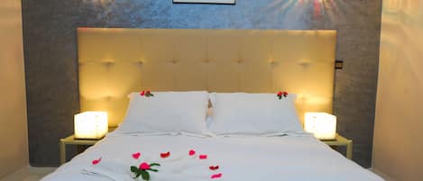 Una cama bien hecha con sábanas blancas y pétalos de rosa, dos lámparas de noche y un cuadro en la pared.
