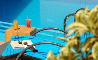 Zona de descanso junto a la piscina con una toalla azul, un libro y dos bebidas sobre un taburete de madera.
