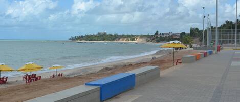 A beachside promenade with a concrete bench, yellow umbrellas, and red chairs along the shore.