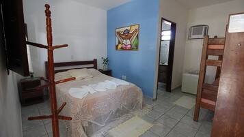Quarto com cama, cadeira de madeira, mesa de cabeceira, mesinha e televisão fixada na parede.