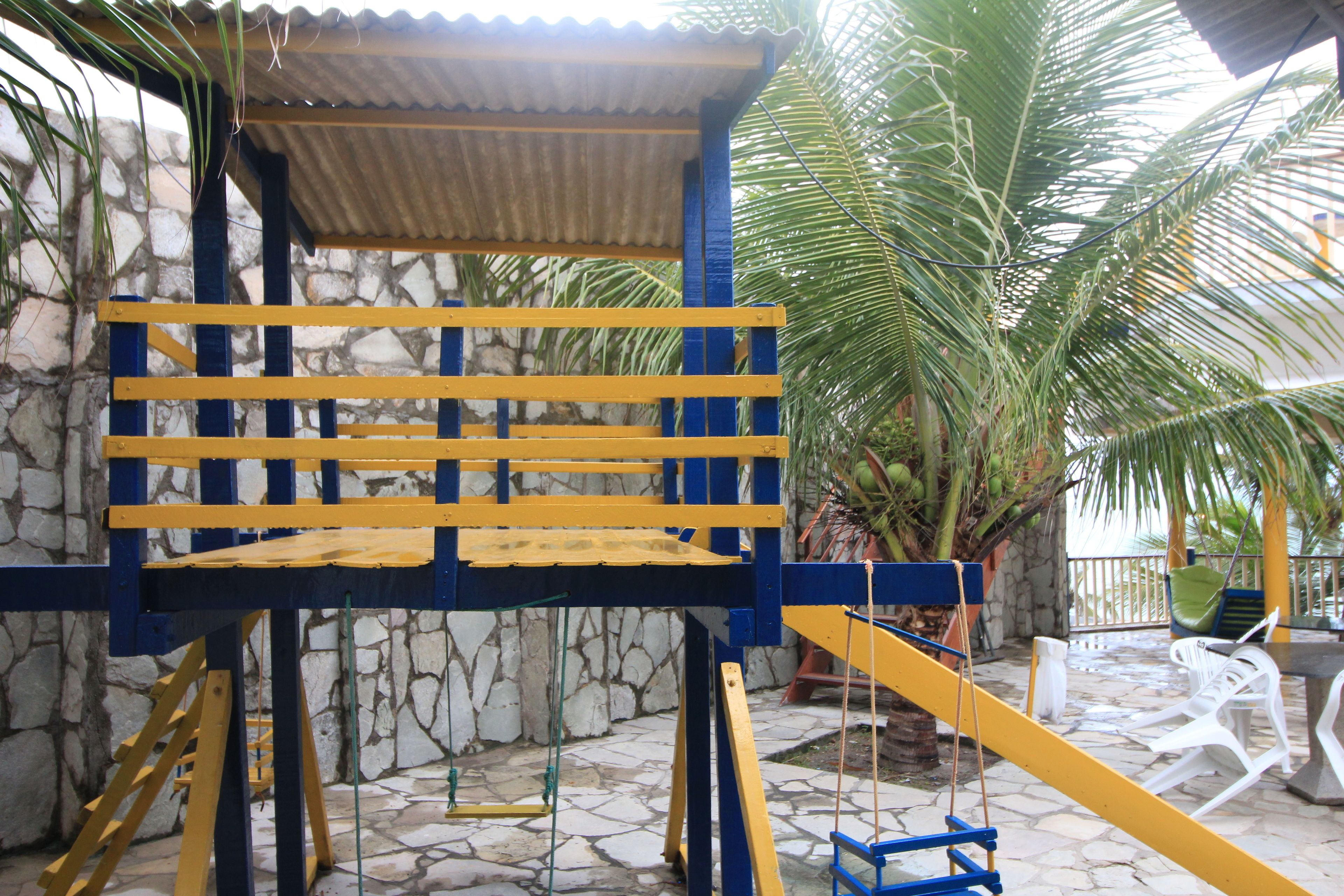 A wooden swing set with yellow and blue colors, a stone wall, and a palm tree in the background.