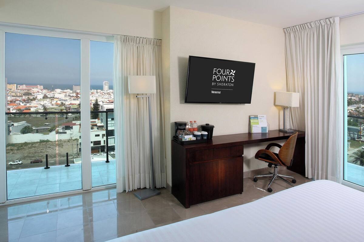Een hotelkamer met een groot raam met uitzicht op de stad, een flatscreen televisie aan de muur, een bureau met stoel, en een bed.