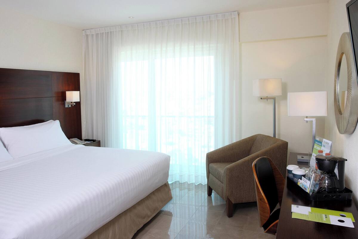 Hotelkamer met een groot bed, een stoel, een bureau met een koffiezetapparaat en een raam met dunne gordijnen.