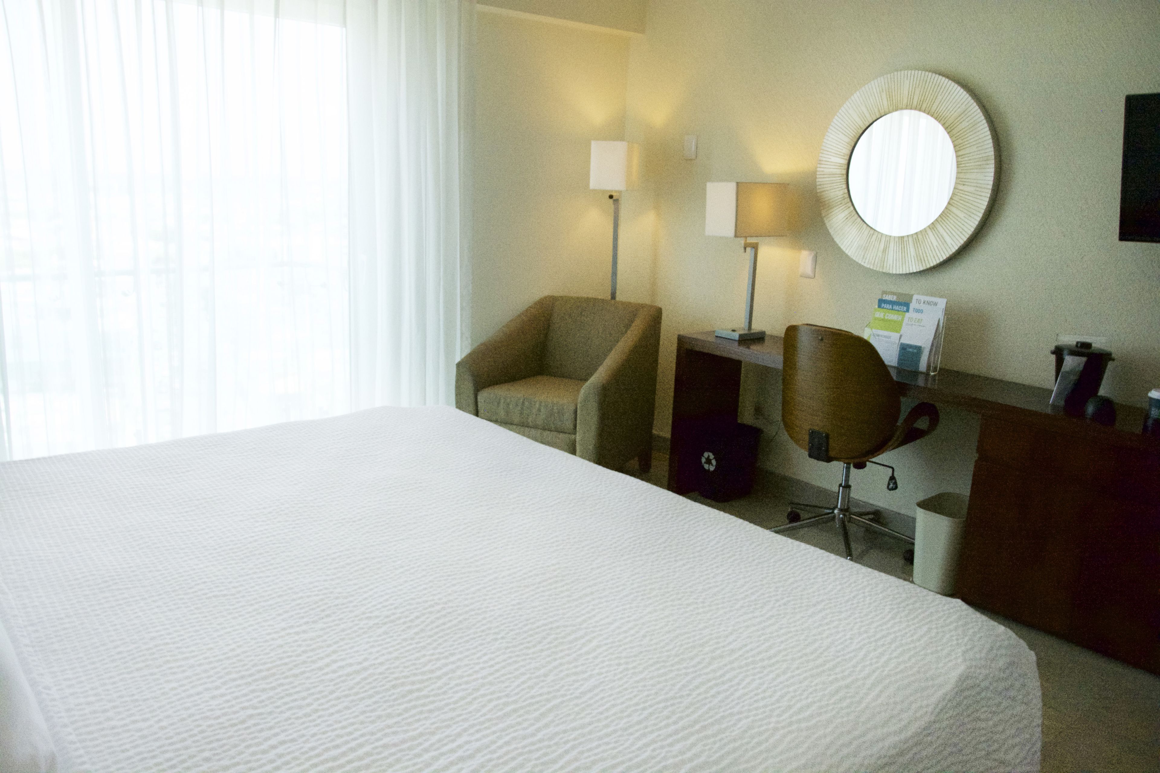 A hotel room with a large bed, two armchairs, a desk with a lamp, a round mirror, and a small trash can.