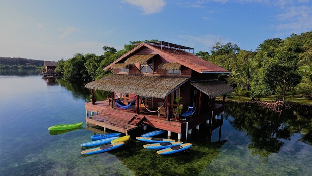 A wooden house with a thatched roof on stilts over clear water, surrounded by greenery. - Casa Del Mar (Tierra Oscura)