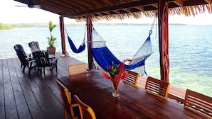 A wooden deck with a table and chairs, a blue hammock, and a view of a body of water. - Casa Del Mar (Tierra Oscura)