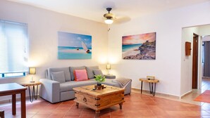 Uma sala de estar com um sofá cinzento, uma mesa de centro de madeira, uma luminária e duas pinturas nas paredes.