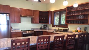A kitchen with wooden cabinets, granite countertops, a large island with bar stools, a refrigerator, stove, and a window with a view of greenery.
