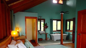 A bedroom with a bed, a bedside table, a lamp, a window, and a bathroom with a sink and bathtub.