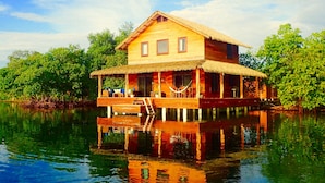 A wooden houseboat with a thatched roof, multiple windows, and a deck with seating.