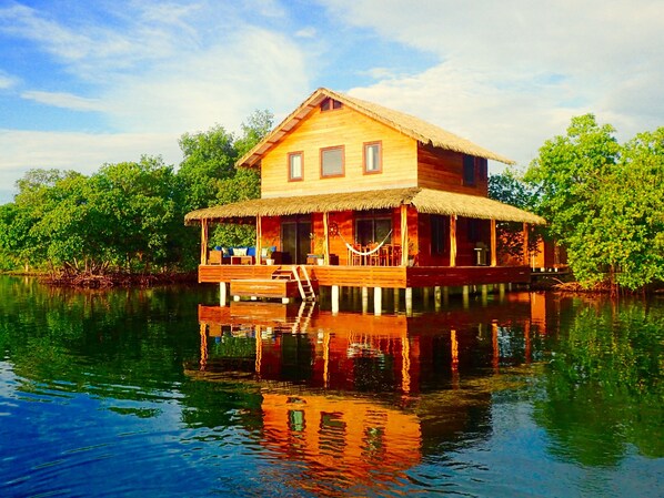 A wooden houseboat with a thatched roof, multiple windows, and a deck with seating. - Casita Del Mar (Tierra Oscura)