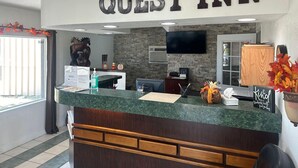 A reception area with a 'QUEST INN' sign, a mounted TV, a wooden cabinet, and a counter with a vase of flowers. - Quest Inn (Weatherford)