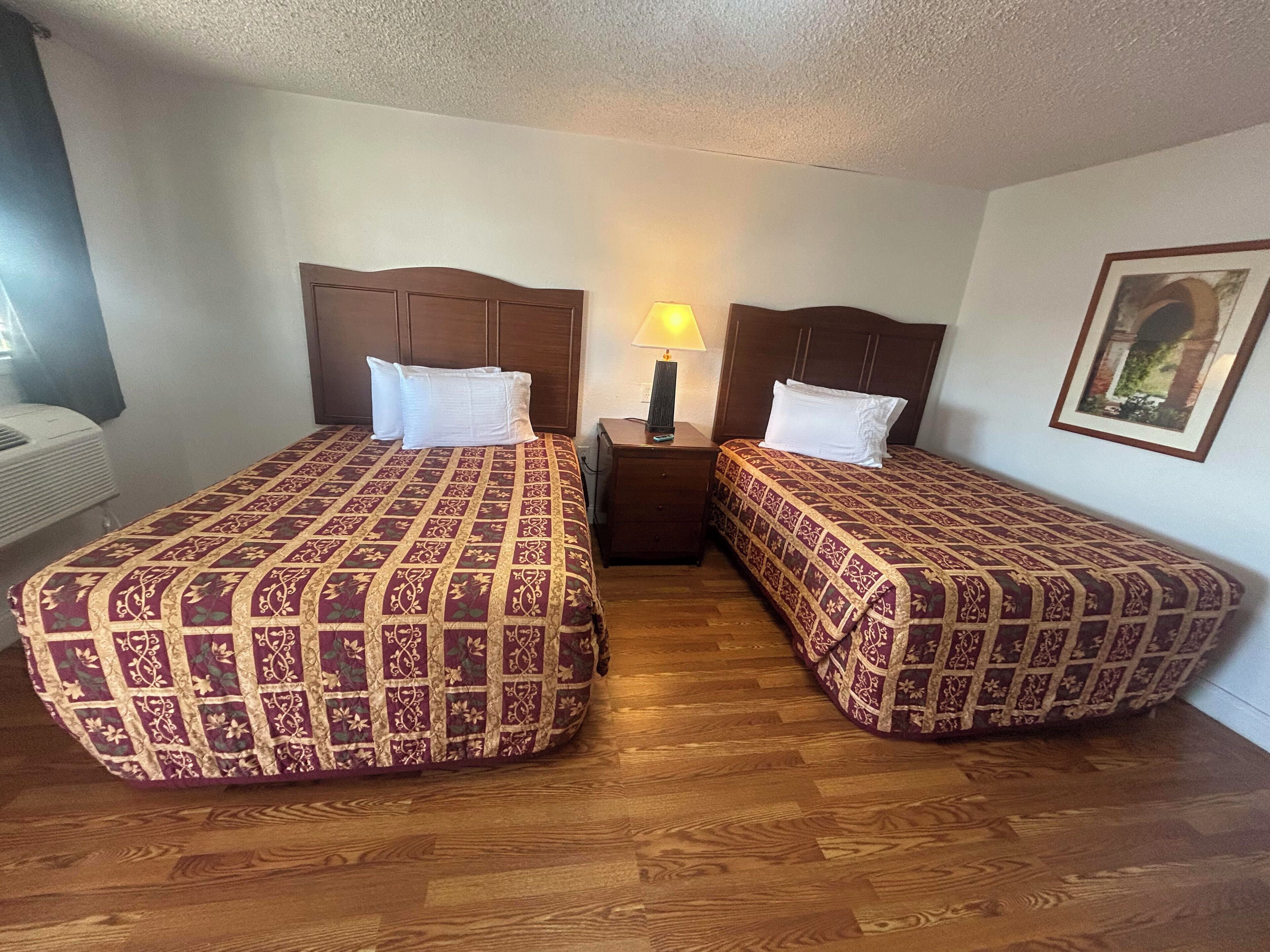A room with two beds, each with a patterned bedspread, a nightstand with a lamp, and a framed picture on the wall.