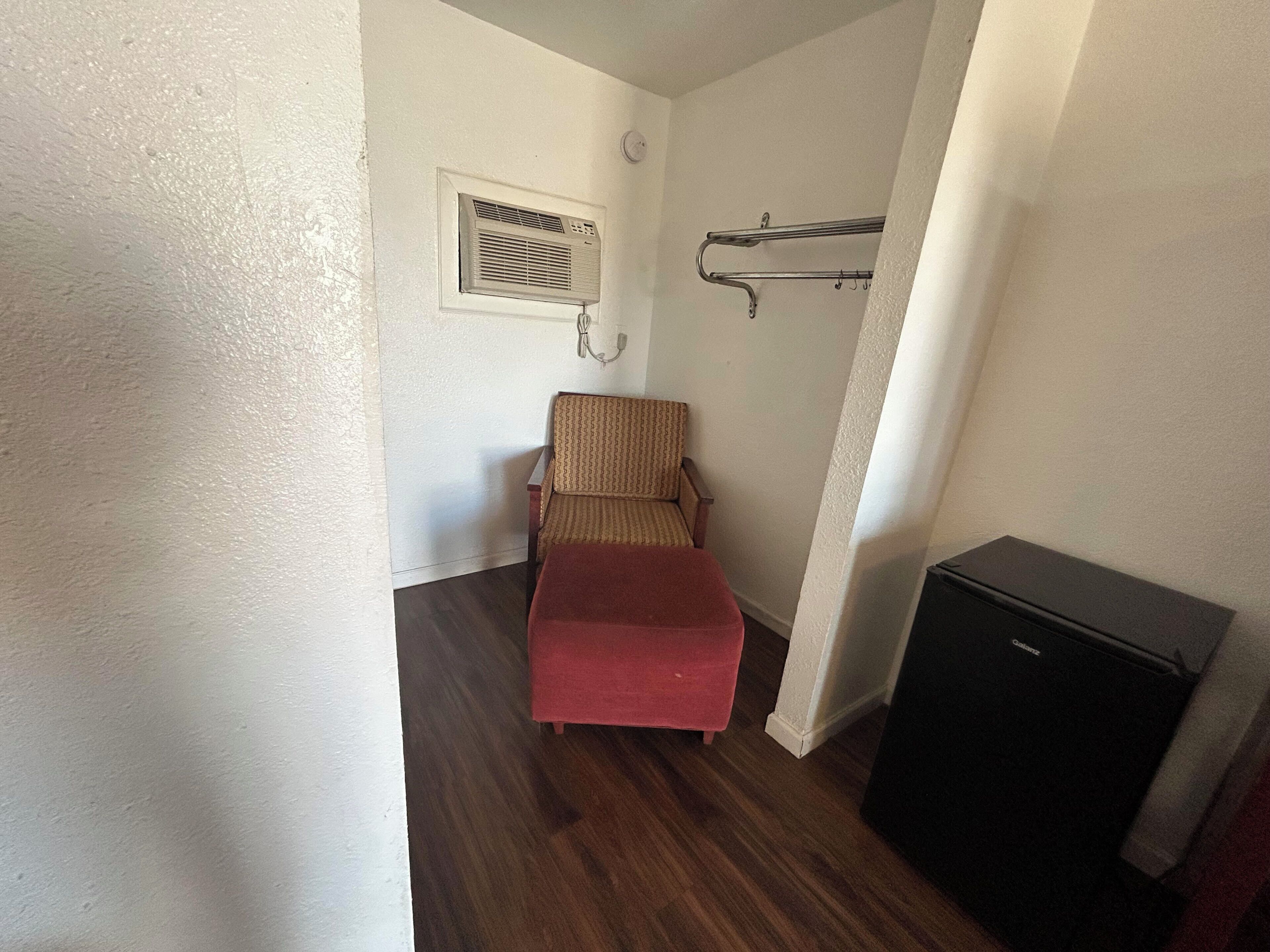 A compact room with a wooden floor, a red sofa, a chair, a small black cabinet, and a wall-mounted air conditioning unit.