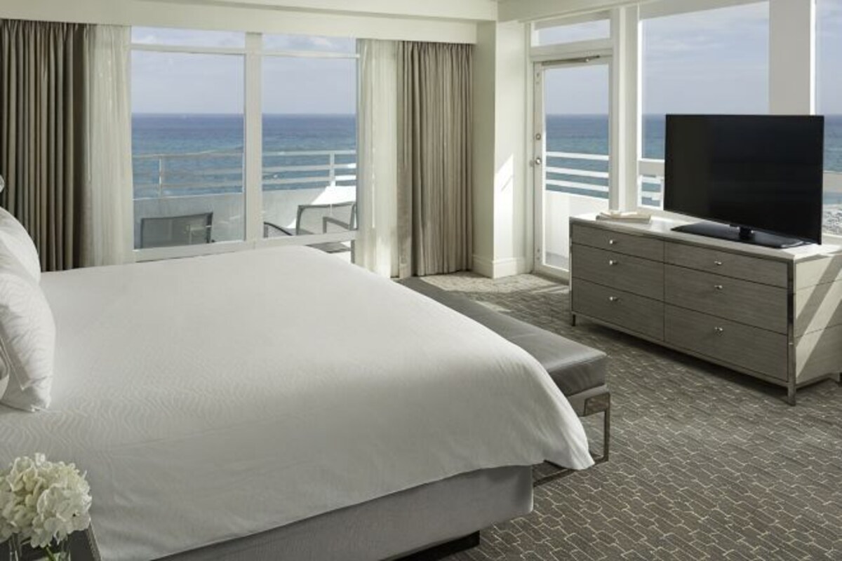 Habitación de hotel con una cama grande, televisor, cómoda y balcón con vista al océano.