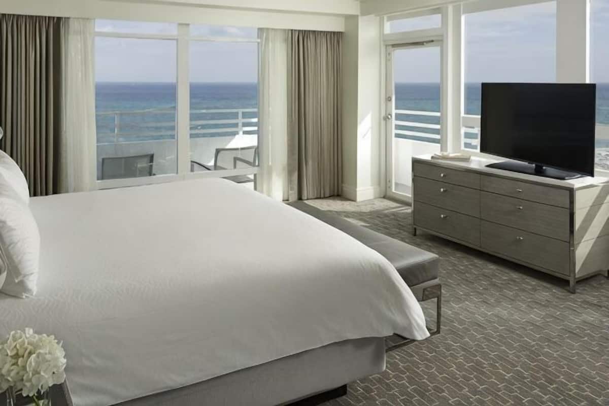 A hotel room with a large bed, a TV, a dresser, and a balcony with a view of the ocean.
