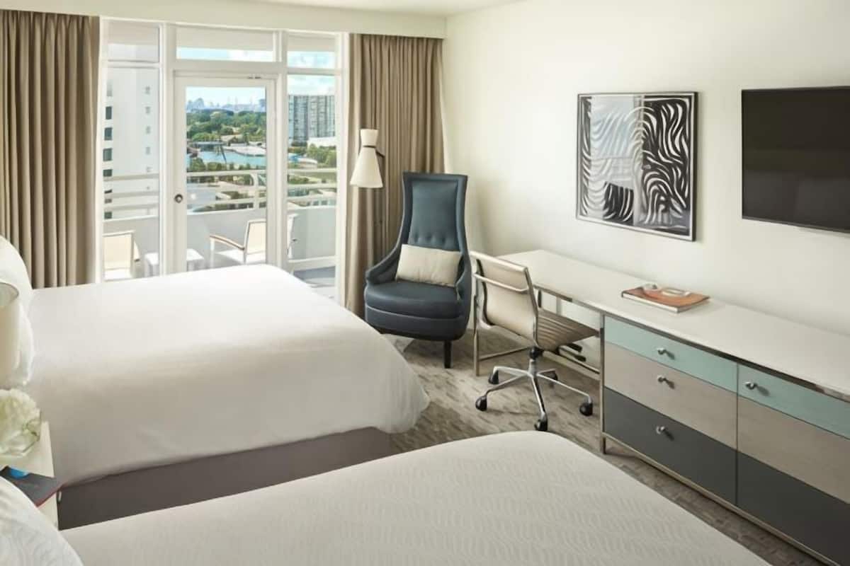 A hotel room with two beds, a desk, a chair, a TV, and a balcony with a view.