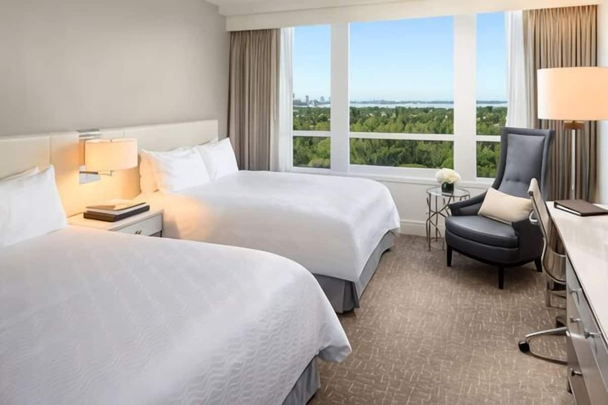 A hotel room with two beds, a desk, a chair, and a view of a cityscape.