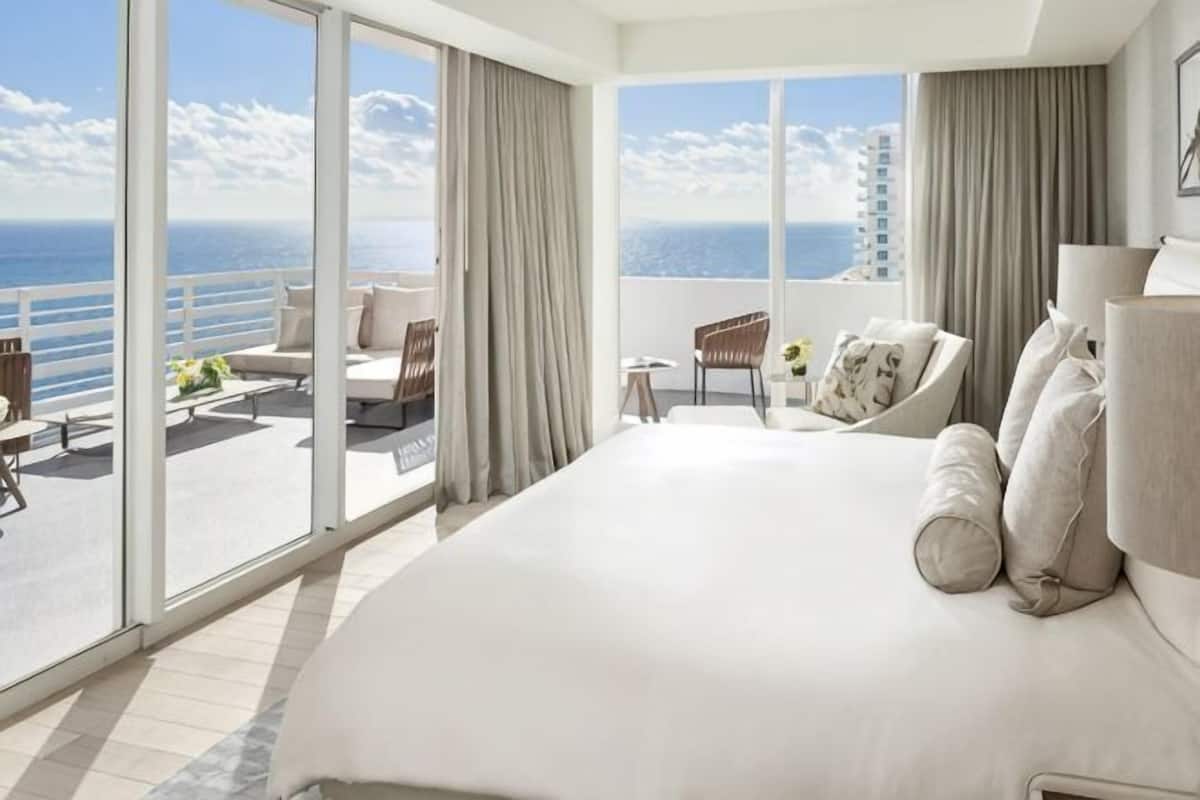 A large bed with a view of the ocean, a balcony with seating, and a large window.