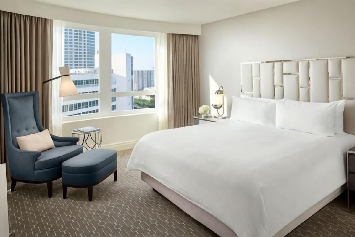 A hotel room with a large bed, a chair, a small table, and a view of the city through the window.